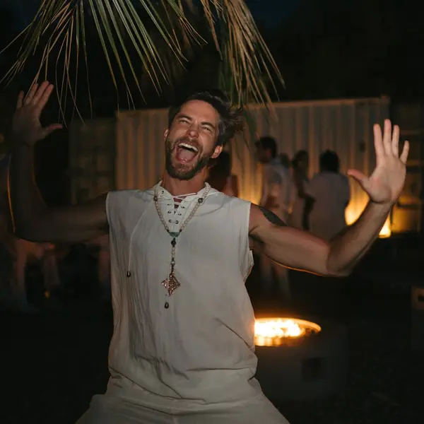 Scott mid-laugh, arms raised, at a night gathering by a fire